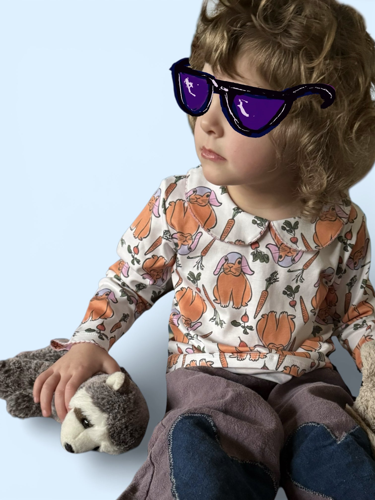 Child wearing a patterned shirt with bunnies and vegetables, holding a stuffed animal, on a light blue background.